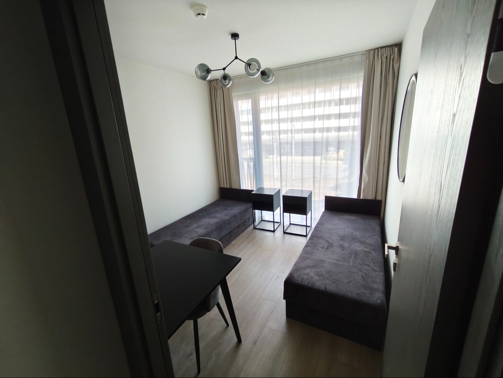 Jasne wnętrze pokoju z dwoma tapczanami, stolikami nocnymi i stołem z krzesłem, widziane z perspektywy otwartych drzwi. Nowoczesny design, neutralna kolorystyka.