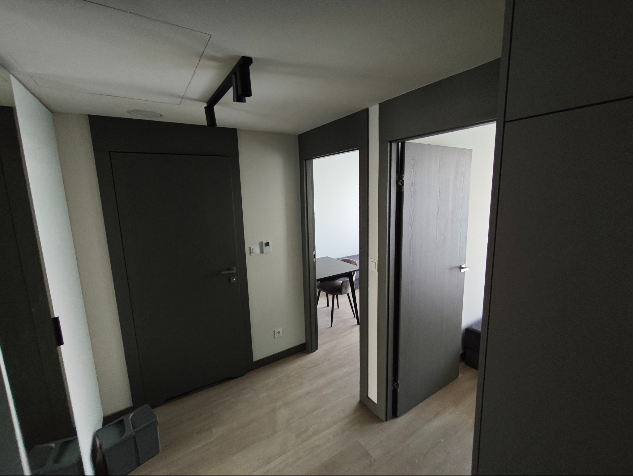 Nowoczesny korytarz z szarymi drzwiami i panelami podłogowymi. Widoczne oświetlenie sufitowe i fragment stołu z krzesłami w tle.