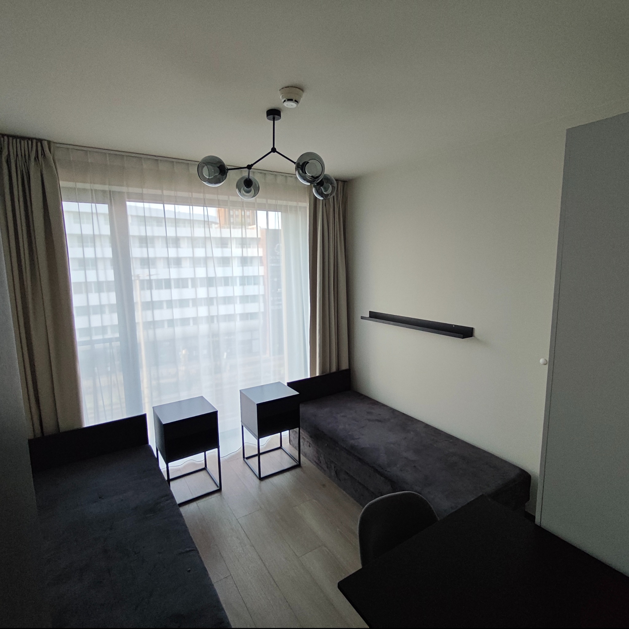 Nowoczesny pokój z dwoma łóżkami, stolikami nocnymi i dużym oknem z widokiem na budynek. Minimalistyczny design, szare odcienie i drewniana podłoga.