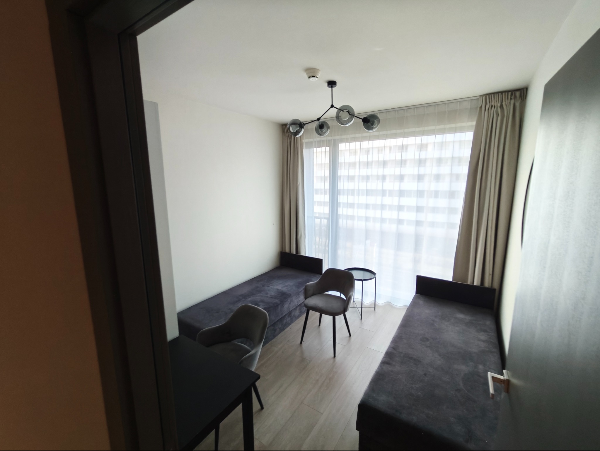 Jasne wnętrze pokoju z dwoma tapczanami, krzesłami i nowoczesną lampą sufitową. Widok przez otwarte drzwi.