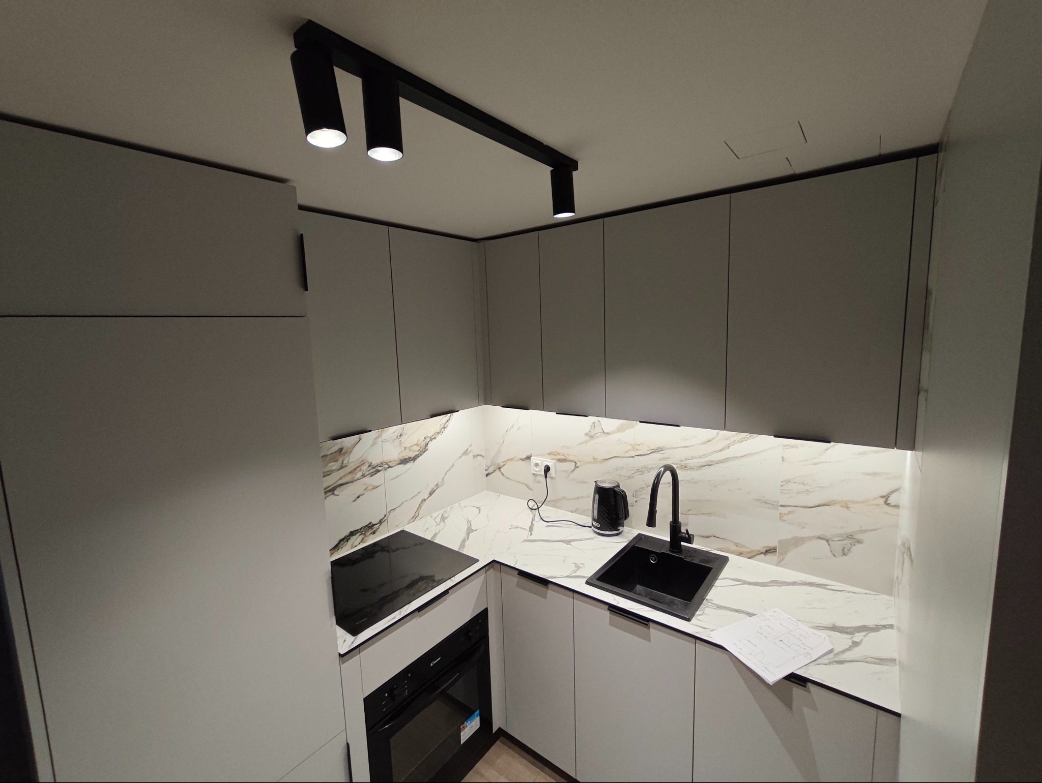 Nowoczesna, szara kuchnia z marmurowym blatem i czarną armaturą. Oświetlenie punktowe LED. Na blacie czajnik i plany kuchni. Minimalistyczny design.