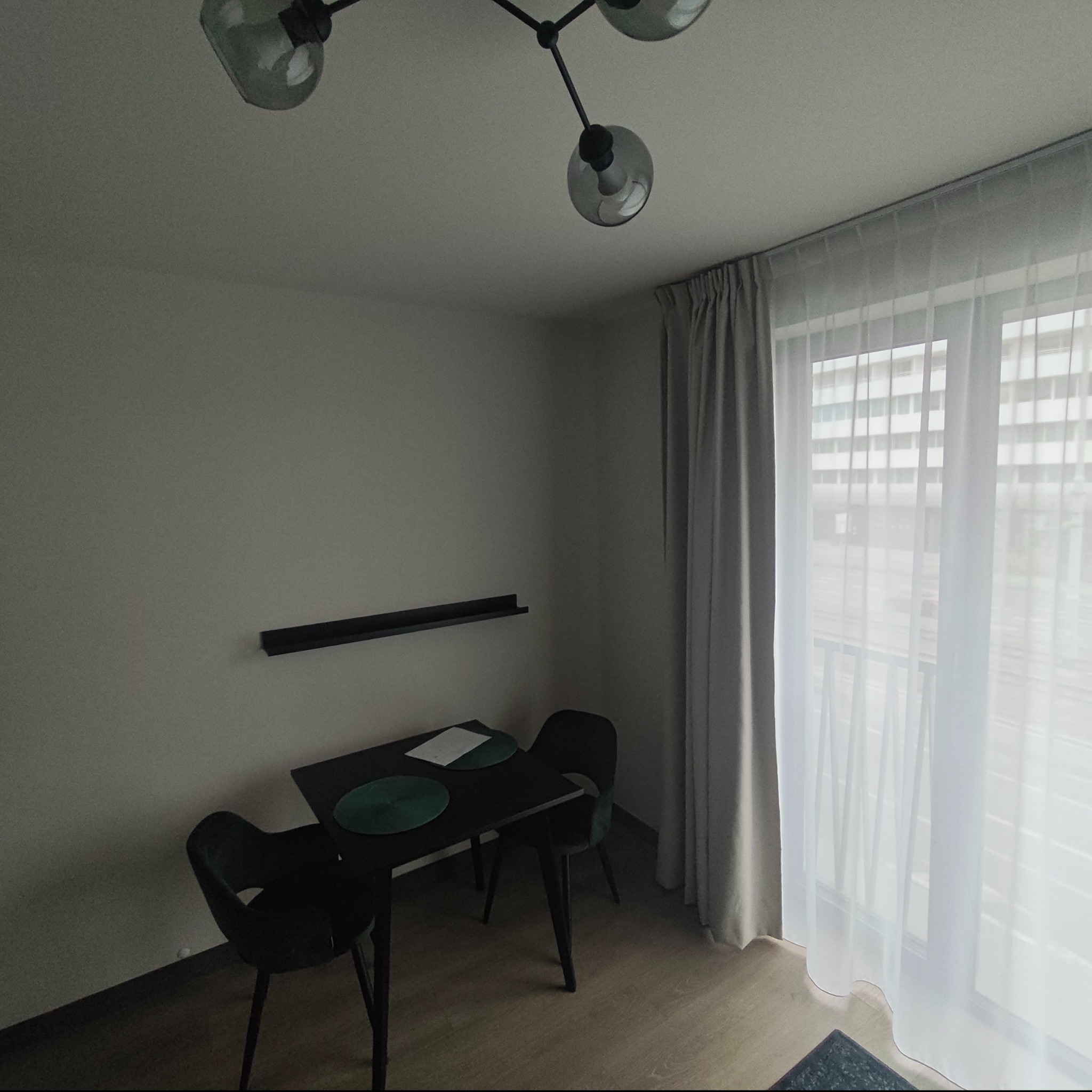 Minimalistyczna jadalnia: czarny stół z krzesłami, półka na ścianie, lampa sufitowa z kulistymi kloszami. Okno z zasłonami wpuszcza dużo światła. Stonowana kolorystyka.