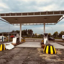 Nowo wybudowana stacja paliw z betonowymi stanowiskami, żółto-czarnymi barierami i zadaszeniem, widoczna w tle biała budka TOI TOI i samochody.