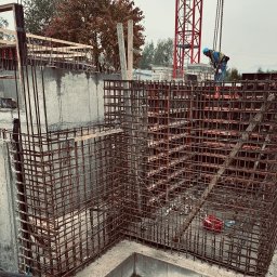 Dominik Milaniuk DOMINŻ - Zbrojenie fundamentów budynku w trakcie budowy z widoczną konstrukcją szalunkową, pracownikiem w kasku i żurawiem w tle.