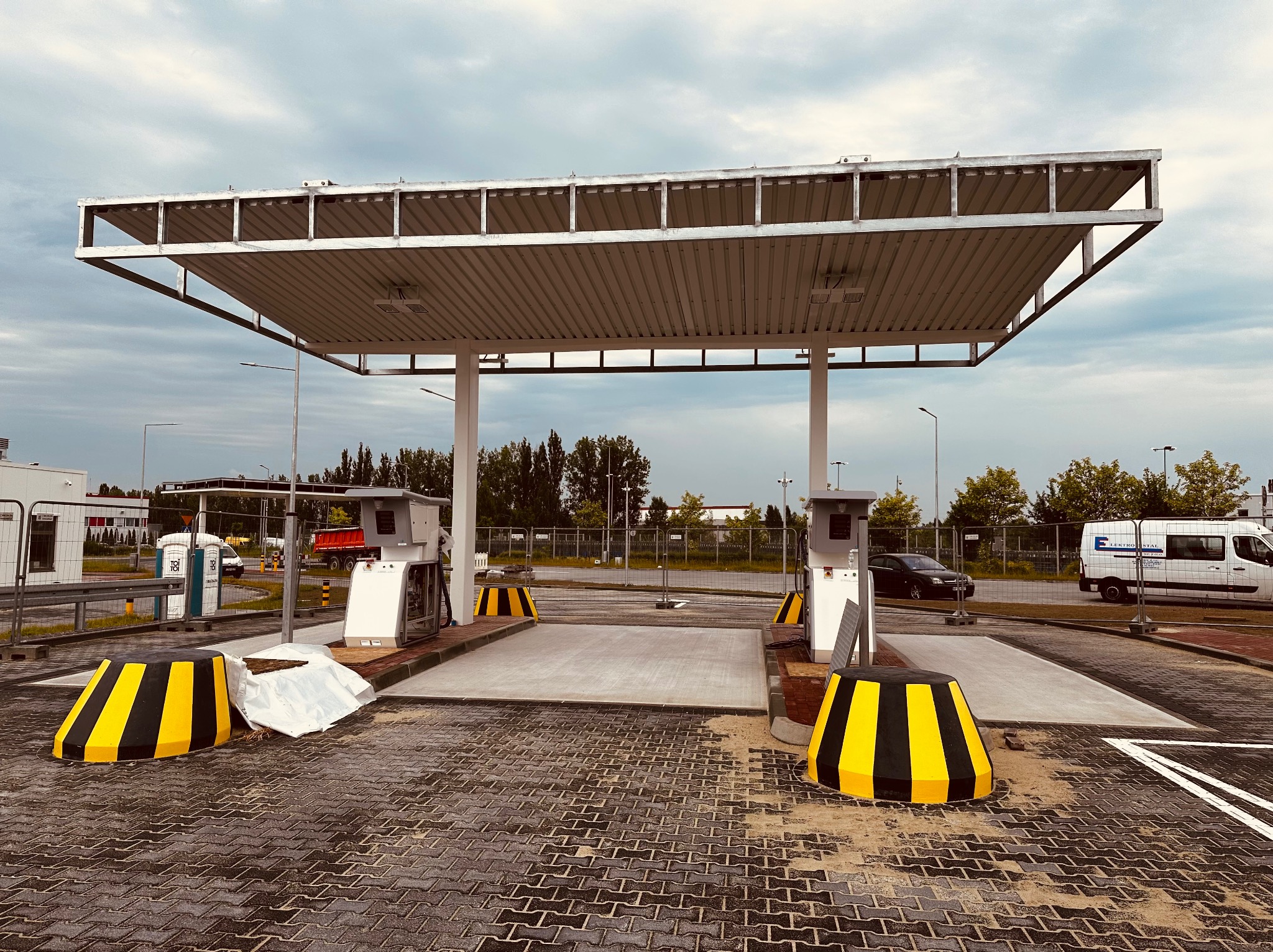 Nowo wybudowana stacja paliw z betonowymi stanowiskami, żółto-czarnymi barierami i zadaszeniem, widoczna w tle biała budka TOI TOI i samochody.