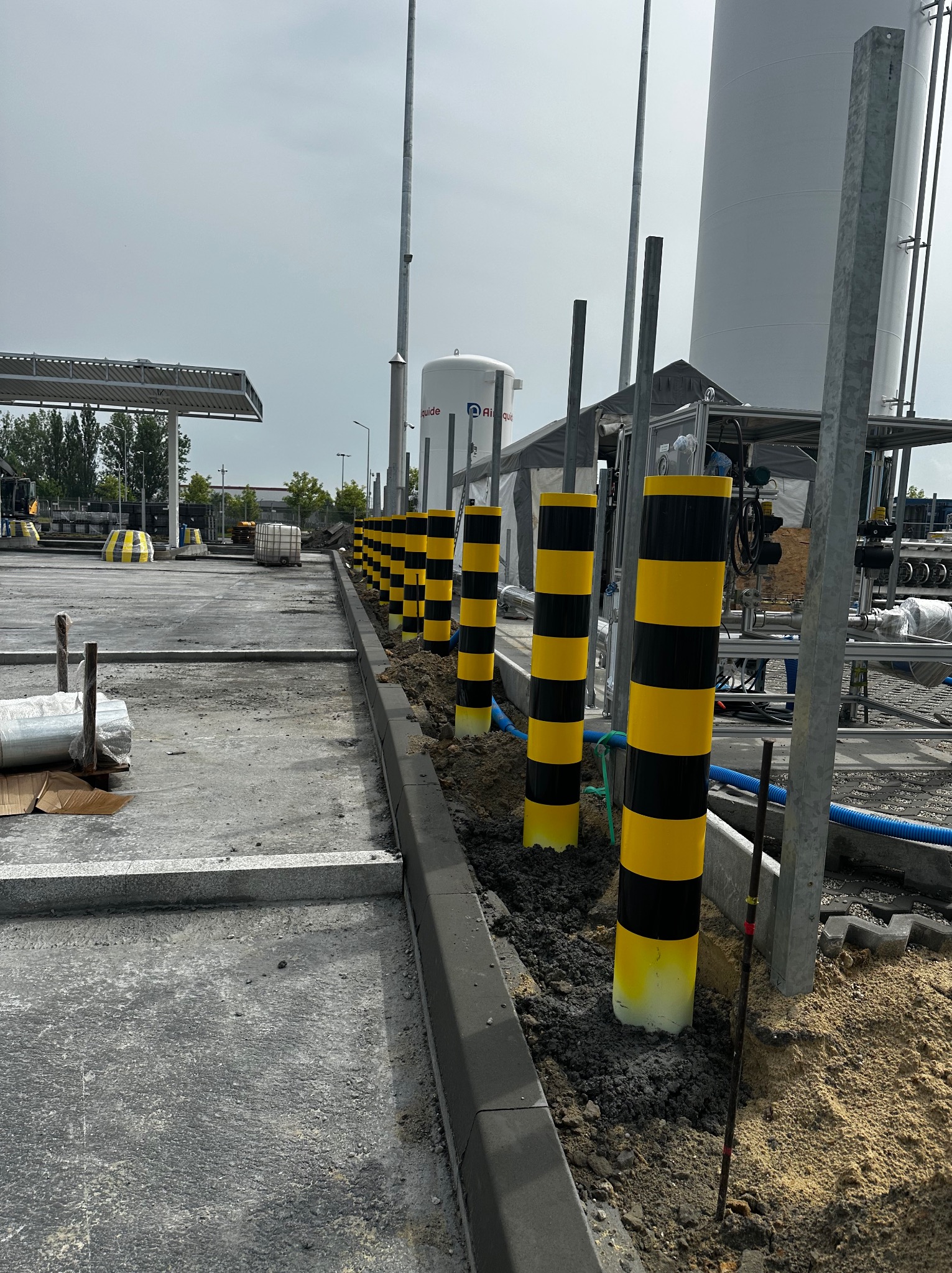 Montaż słupków ochronnych, pomalowanych w żółto-czarne pasy, wzdłuż krawężnika na terenie przemysłowym, z widocznymi zbiornikami i instalacjami w tle.