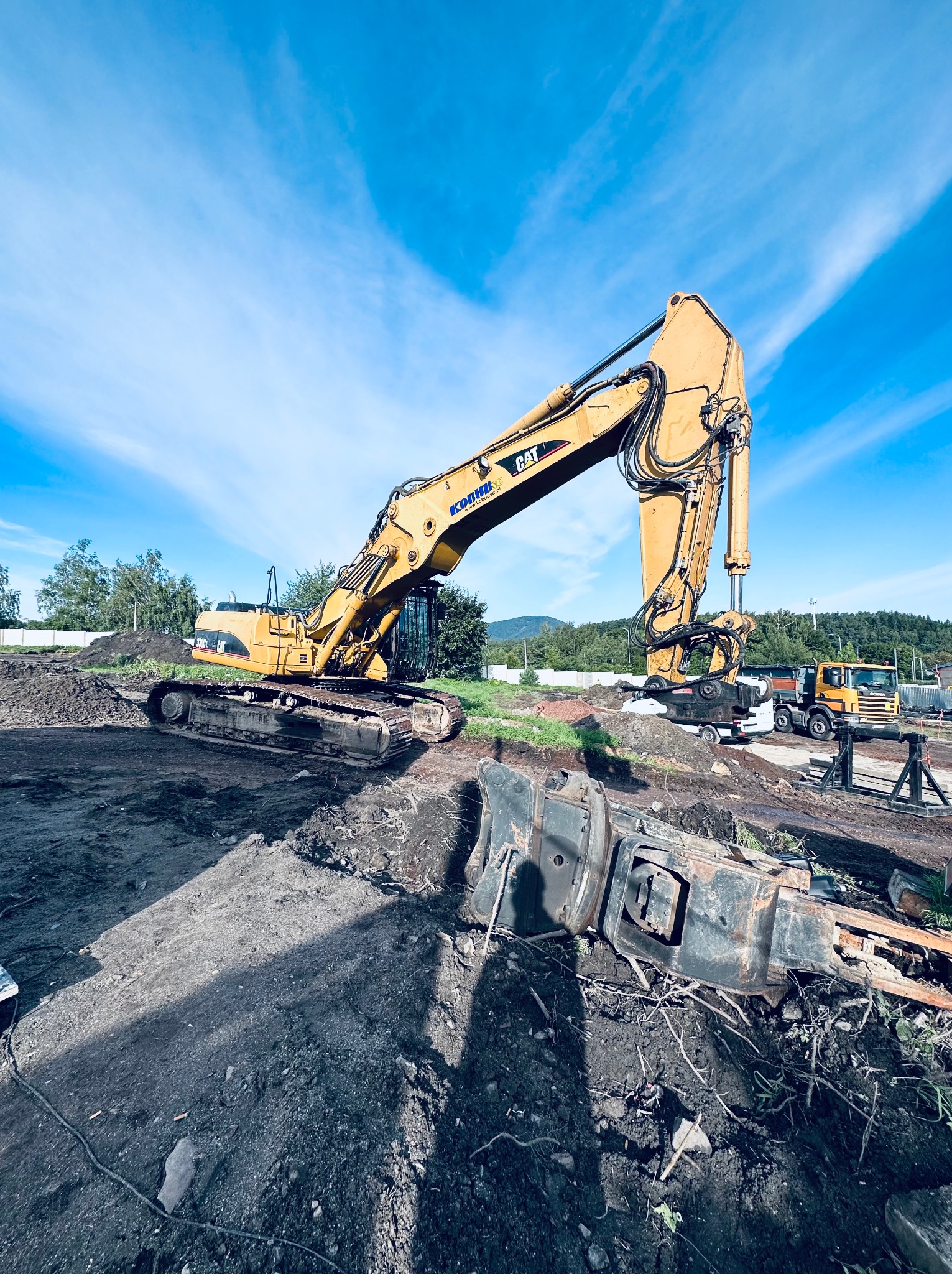 Żółta koparka CAT z wysięgnikiem w górze na placu budowy, z fragmentami rozebranego sprzętu w pierwszym planie, widoczne ciężarówki w tle i błękitne niebo z chmurami.