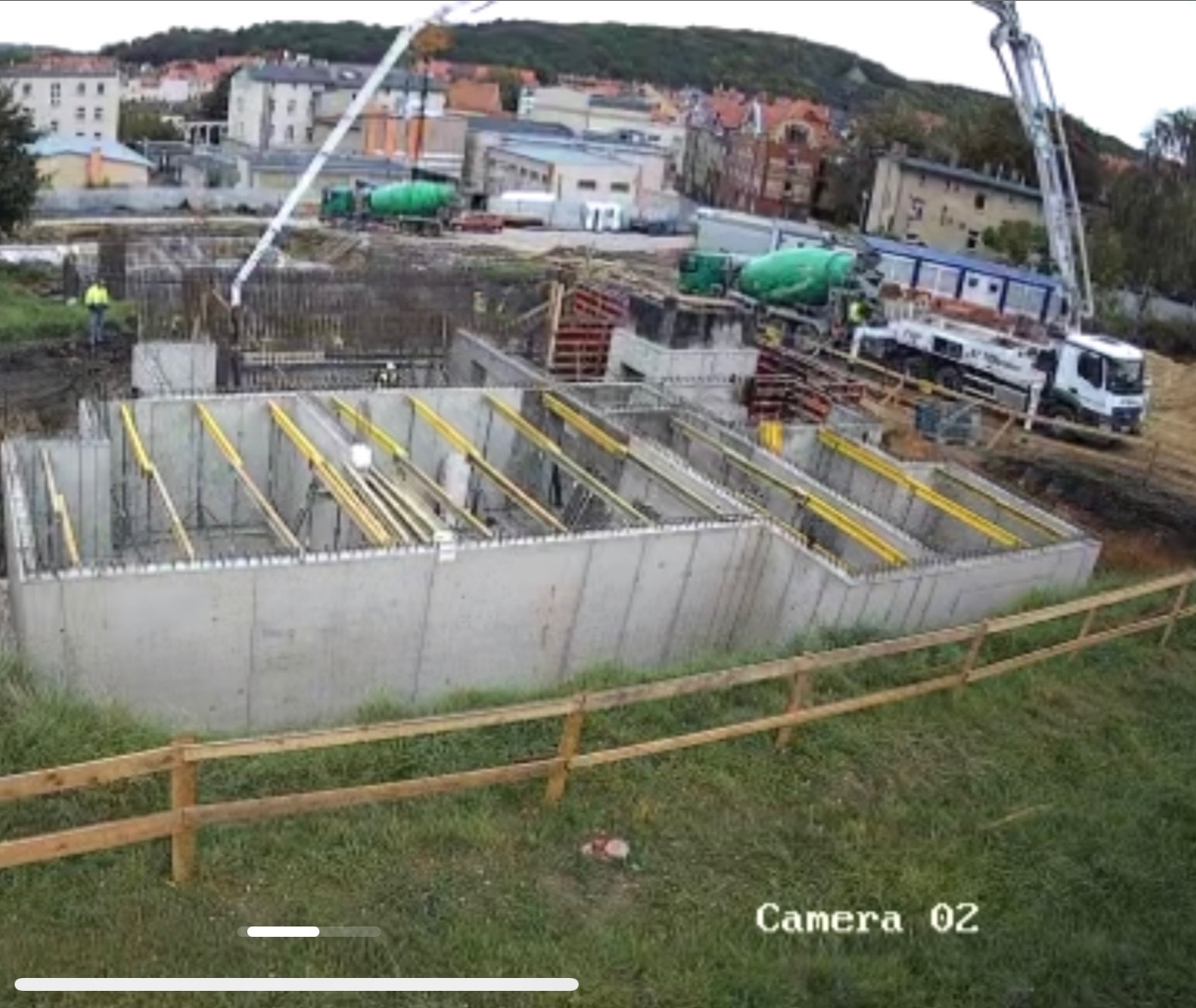 Betonowy fundament budynku w trakcie wylewania, widok z kamery przemysłowej, z betonomieszarką i pompą do betonu w tle.