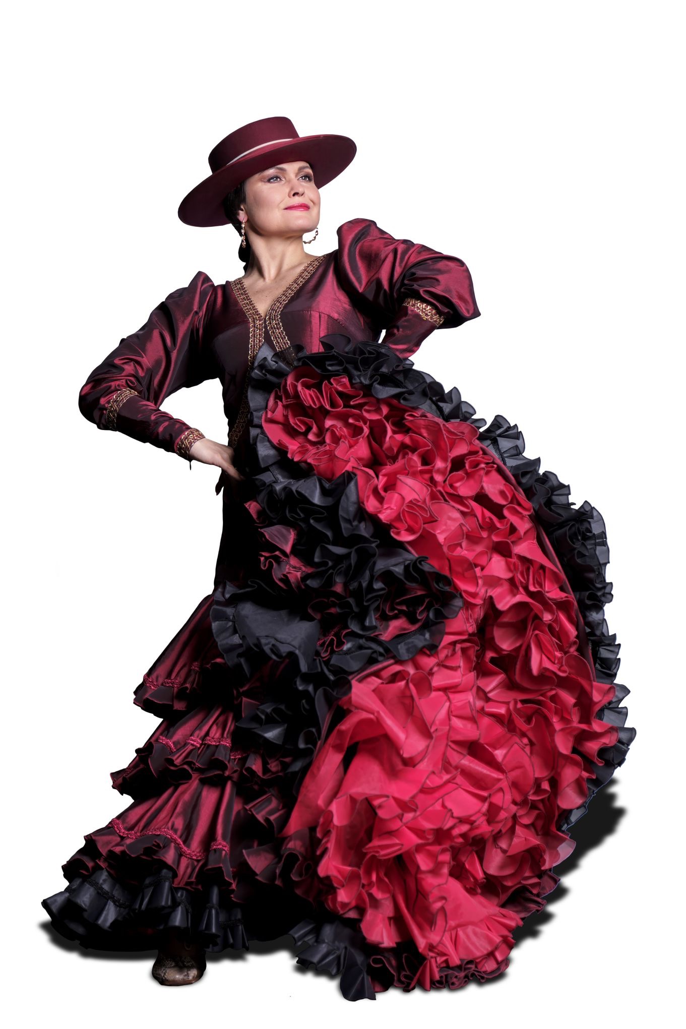 Kobieta w czerwono-czarnej sukni flamenco z falbanami i kapeluszu, pozująca na białym tle, z ręką na biodrze i uniesioną głową.