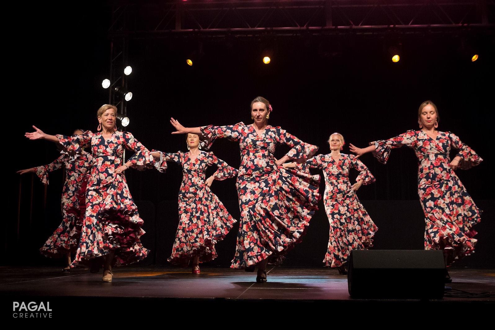 Zespół tancerek flamenco w kwiecistych, falbaniastych sukniach prezentuje układ na scenie oświetlonej reflektorami.