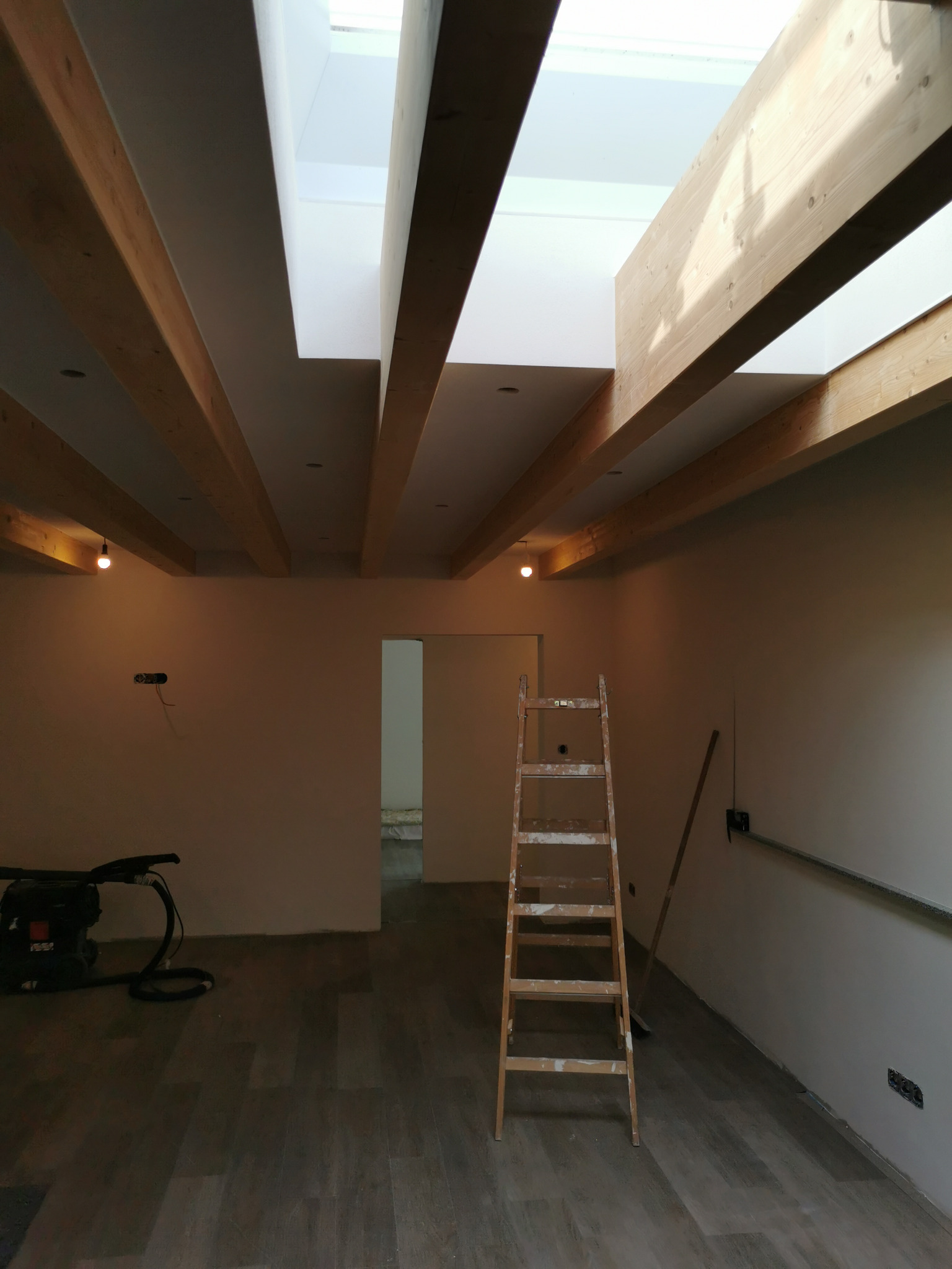 Wnętrze w trakcie prac wykończeniowych: drewniane belki stropowe, sufit z oknem dachowym, drabina malarska, podłoga wyłożona panelami, ściany przygotowane do malowania, widoczny sprzęt budowlany.