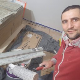 Oleg FENYAK - Mężczyzna w czerwonej bluzie, z wałkiem malarskim w dłoni, robiący selfie na tle przygotowanych płytek podłogowych i narzędzi malarskich.