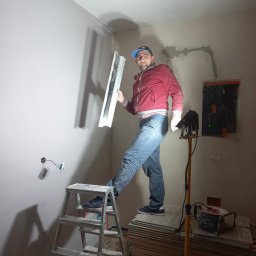 Oleg FENYAK - Mężczyzna na drabinie trzymający szpachelkę na tle świeżo wyszpachlowanej ściany z widocznymi kablami elektrycznymi i lampą oświetleniową.