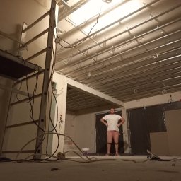 Oleg FENYAK - Wnętrze w trakcie prac wykończeniowych, widoczne metalowe profile podwieszanego sufitu, drabina, instalacje elektryczne i mężczyzna w centralnej części kadru.