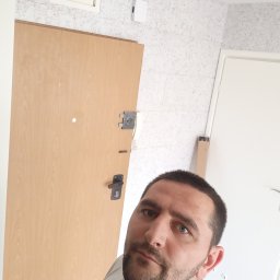 Oleg FENYAK - Mężczyzna w trakcie remontu korytarza z drewnianymi drzwiami wejściowymi i wykończonymi ścianami imitującymi marmur, widoczna skrzynka z bezpiecznikami.