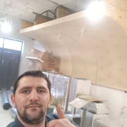 Oleg FENYAK - Mężczyzna robi selfie na tle budowanej konstrukcji z płyty OSB, prawdopodobnie sufitu podwieszanego o łukowym kształcie, widoczne materiały izolacyjne i wentylacyjne.