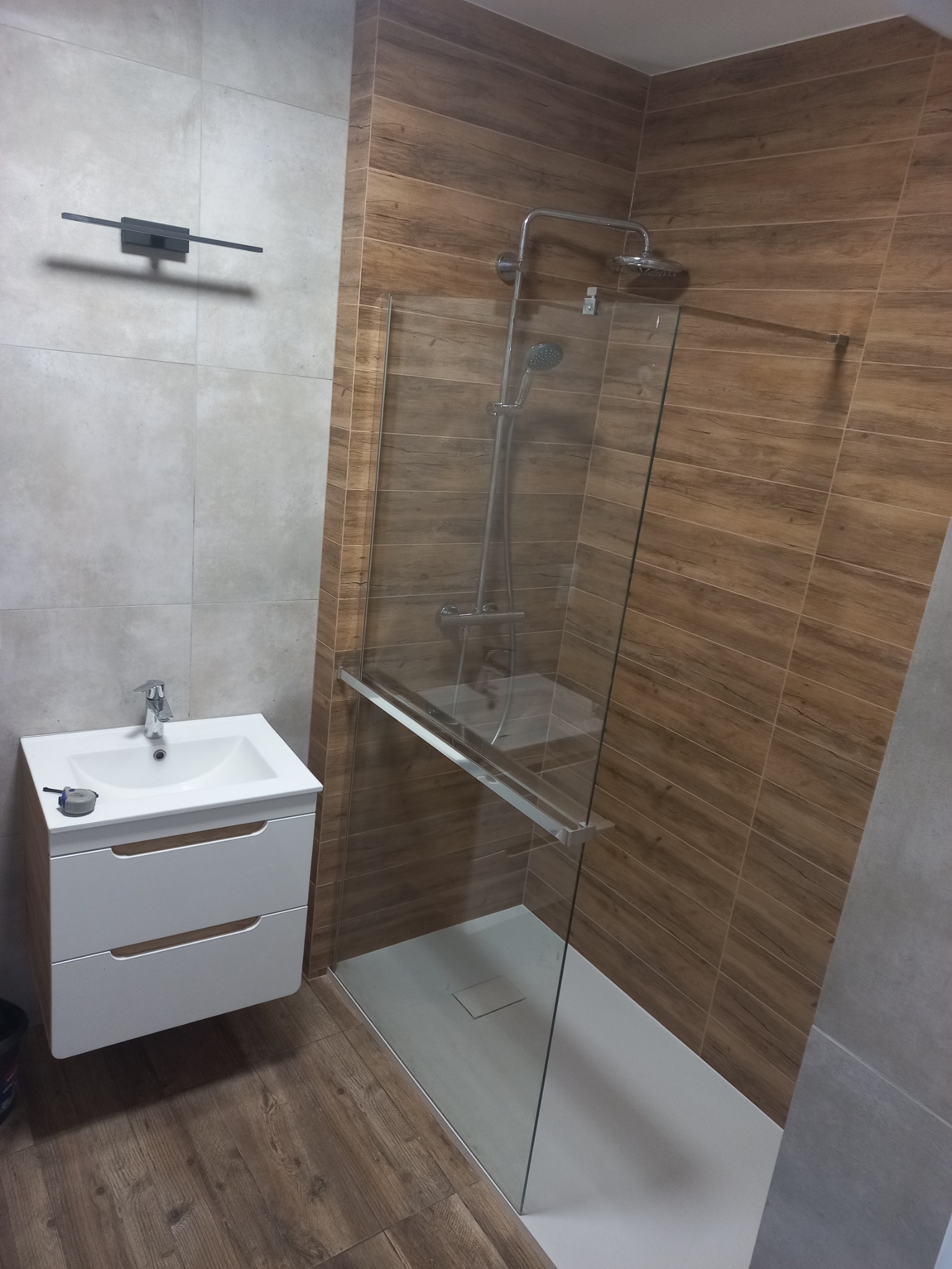 Nowoczesna łazienka z kabiną prysznicową typu walk-in, umywalką z szafką w kolorze białym i płytkami imitującymi drewno na ścianach.