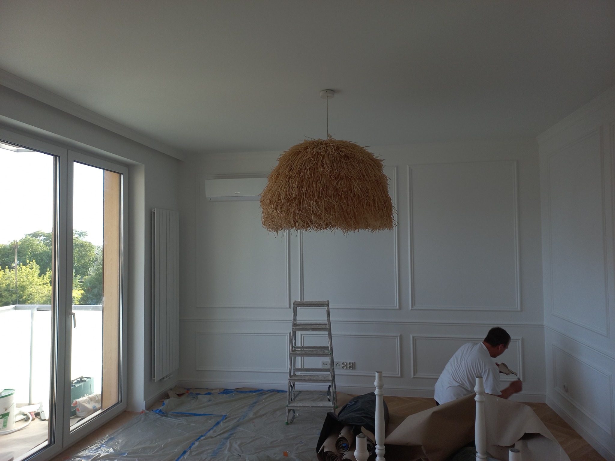Pracownik maluje ścianę z dekoracyjnymi listwami w jasnym wnętrzu, podłoga zabezpieczona folią, widoczna drabina i nietypowa lampa ze słomy