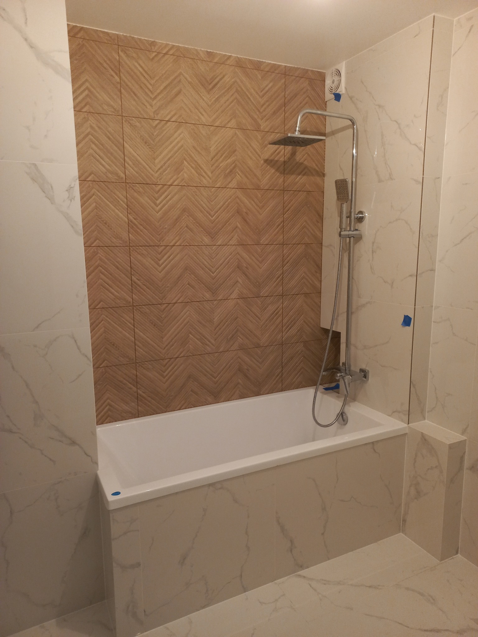 Wykończona łazienka z wanną obudowaną marmurem, brązowe płytki ułożone w jodełkę na ścianie, bateria prysznicowa z deszczownicą i słuchawką, wentylator w rogu.