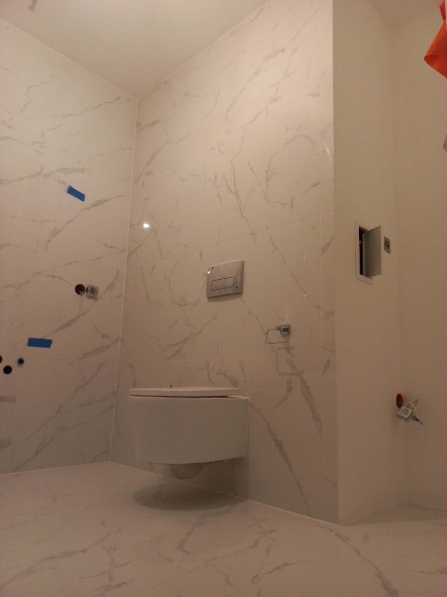 Nowoczesna łazienka z białymi marmurkowymi płytkami, podwieszaną toaletą i elementami instalacji elektrycznej w trakcie montażu.