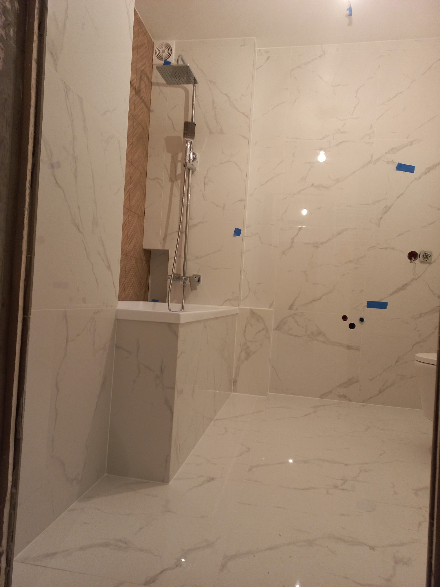 Nowoczesna łazienka w trakcie wykańczania, białe marmurowe płytki na ścianach i podłodze, widoczne niebieskie taśmy malarskie, zamontowana armatura prysznicowa i częściowo zabudowana wanna.