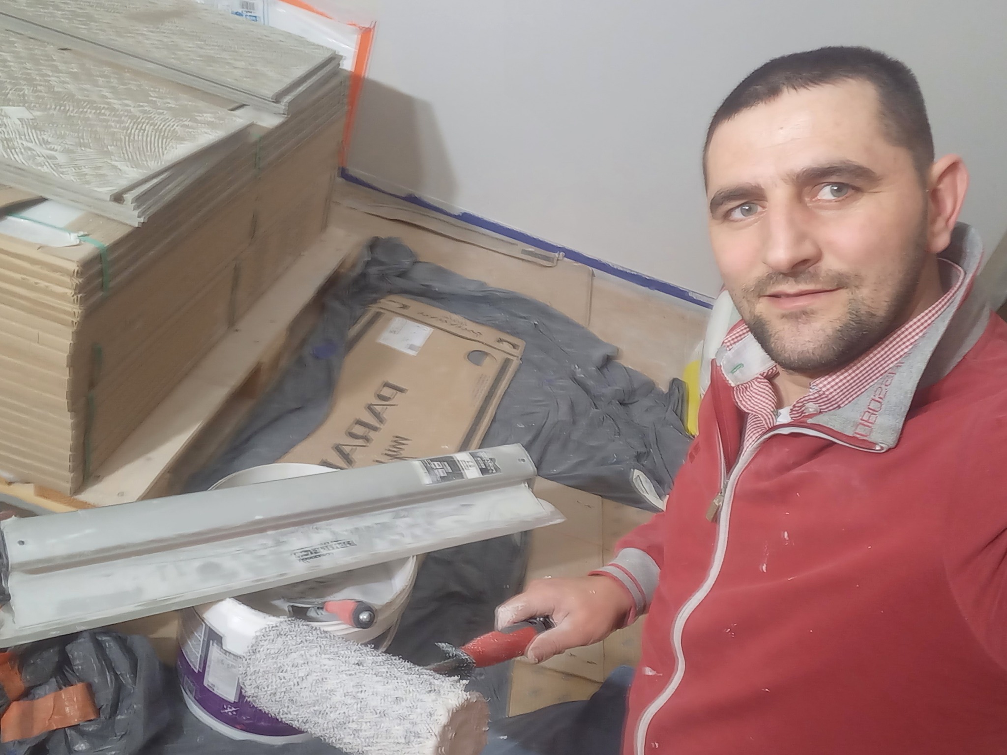 Mężczyzna w czerwonej bluzie, z wałkiem malarskim w dłoni, robiący selfie na tle przygotowanych płytek podłogowych i narzędzi malarskich.