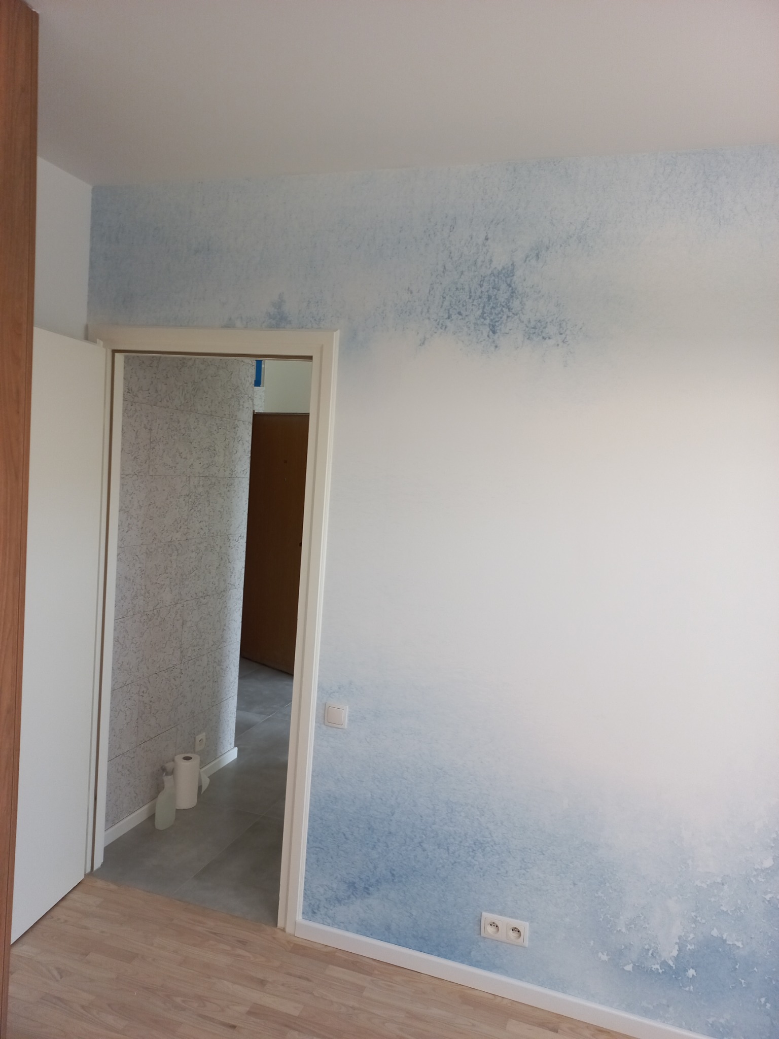 Pokój po malowaniu z efektem ombre w odcieniach błękitu, widoczne gniazdka elektryczne i fragment łazienki z szarą glazurą w tle.