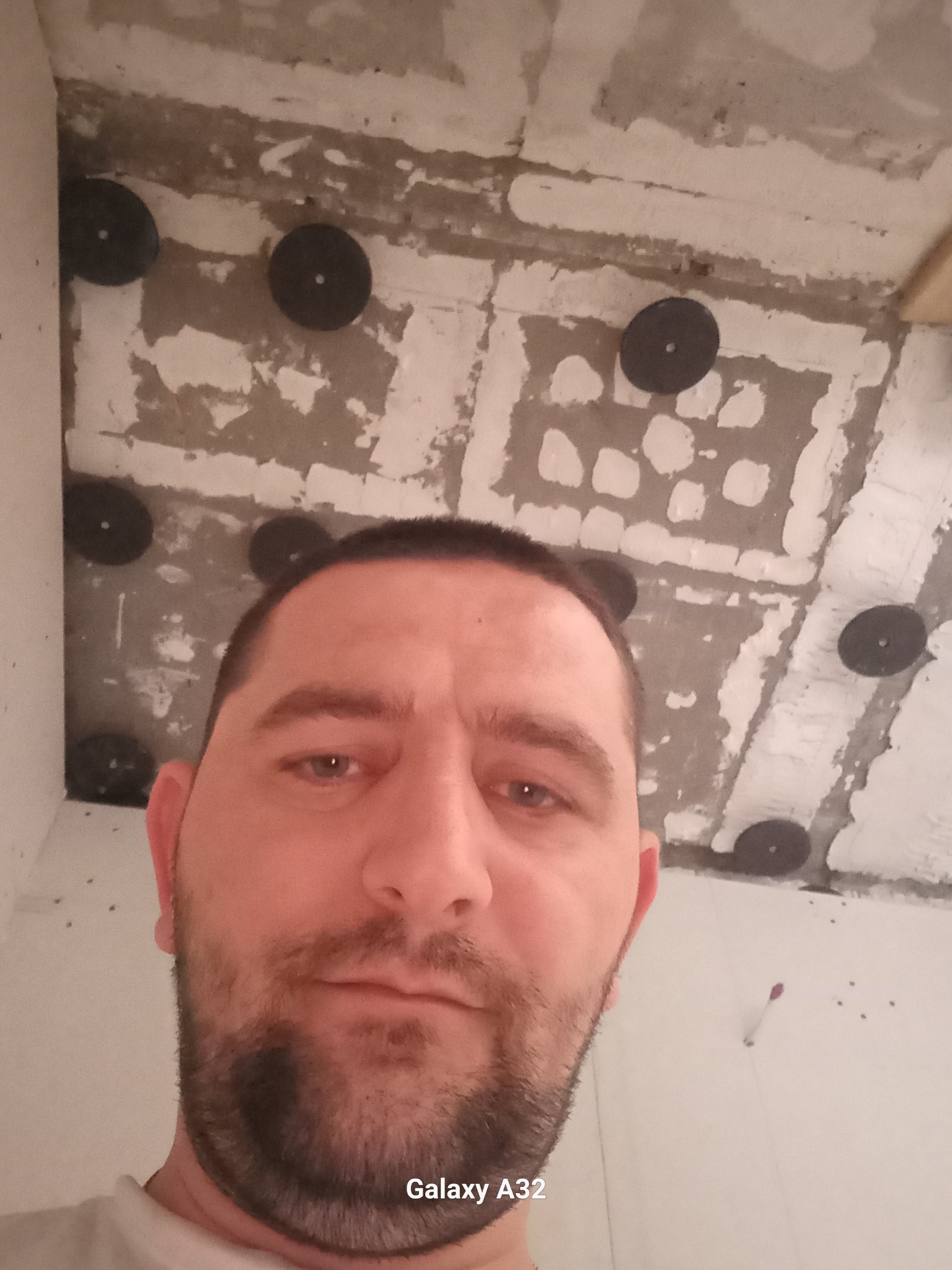 Mężczyzna robiący selfie na tle surowego sufitu z widocznymi mocowaniami do płyt kartonowo-gipsowych, etap przygotowania do prac wykończeniowych.