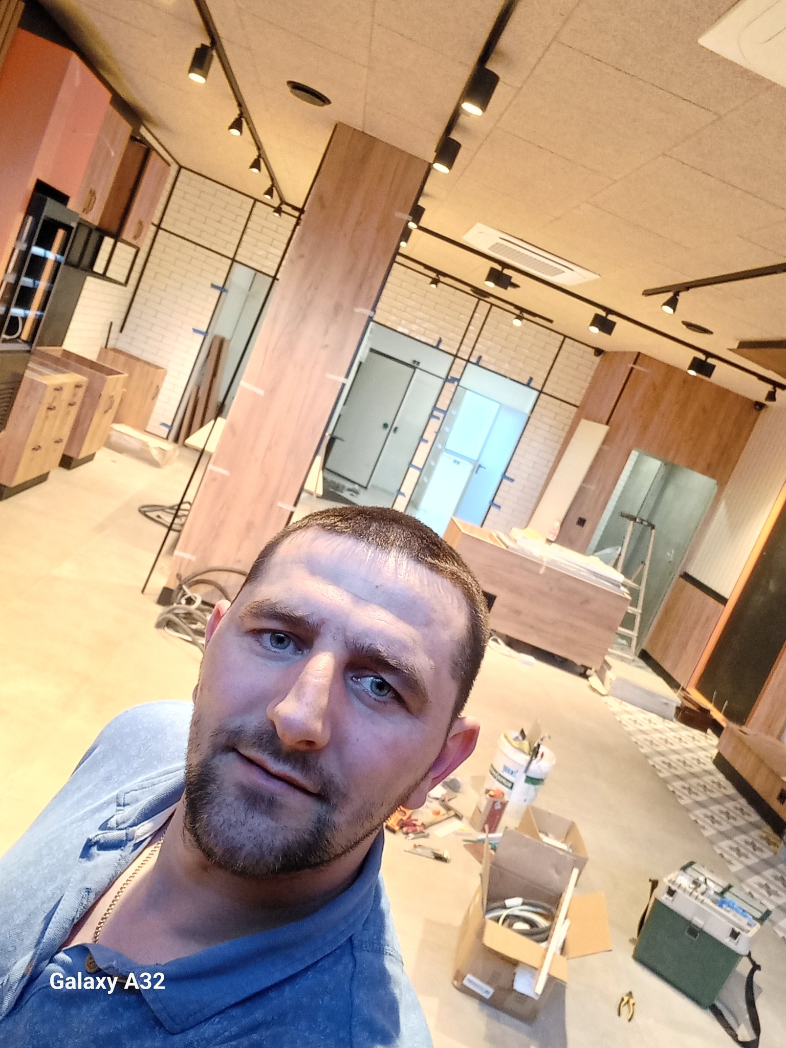 Autoportret wykonawcy na tle wnętrza sklepu w trakcie wykańczania, widoczne białe cegiełki na ścianach, drewniane elementy zabudowy i sufit z oświetleniem szynowym.