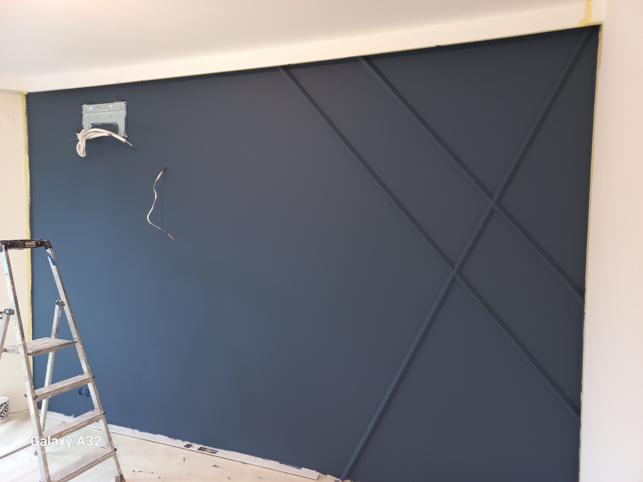 Ściana w kolorze granatowym z geometrycznymi listwami dekoracyjnymi, widoczna drabina malarska i wystające kable od klimatyzacji w trakcie prac wykończeniowych.