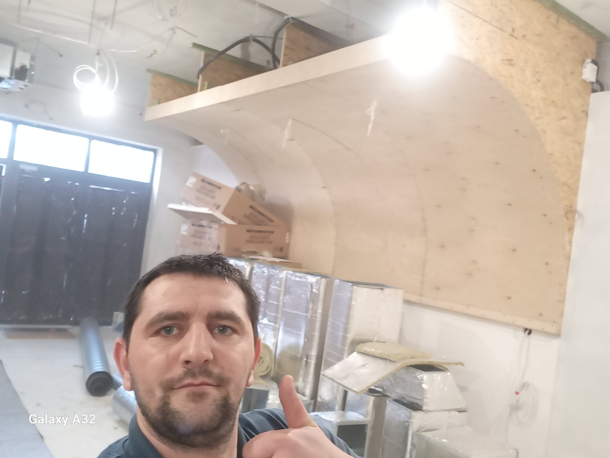 Mężczyzna robi selfie na tle budowanej konstrukcji z płyty OSB, prawdopodobnie sufitu podwieszanego o łukowym kształcie, widoczne materiały izolacyjne i wentylacyjne.