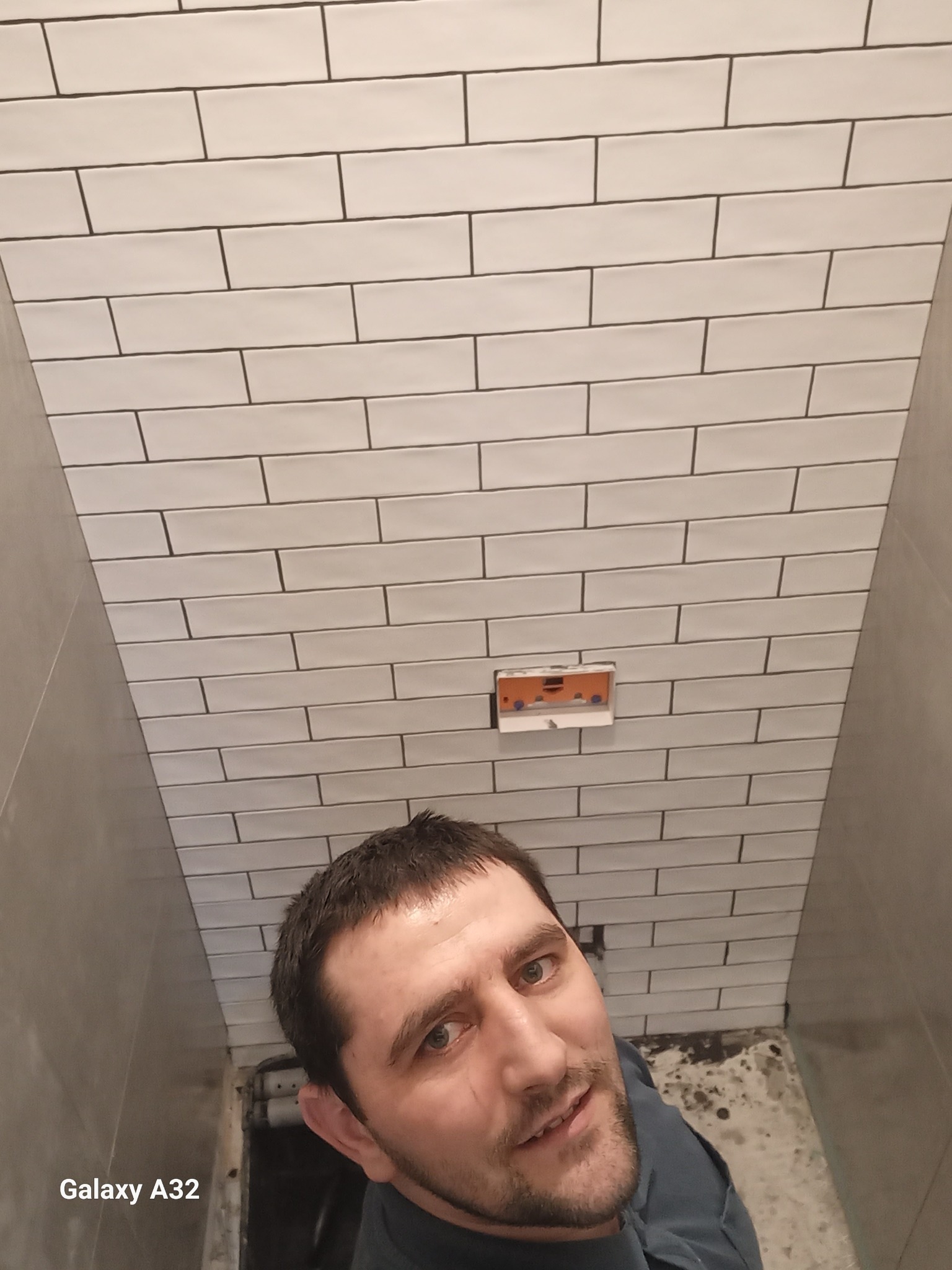 Wnętrze łazienki w trakcie remontu: białe, prostokątne płytki ułożone na ścianie, widoczna puszka elektryczna i fragment mężczyzny robiącego zdjęcie.