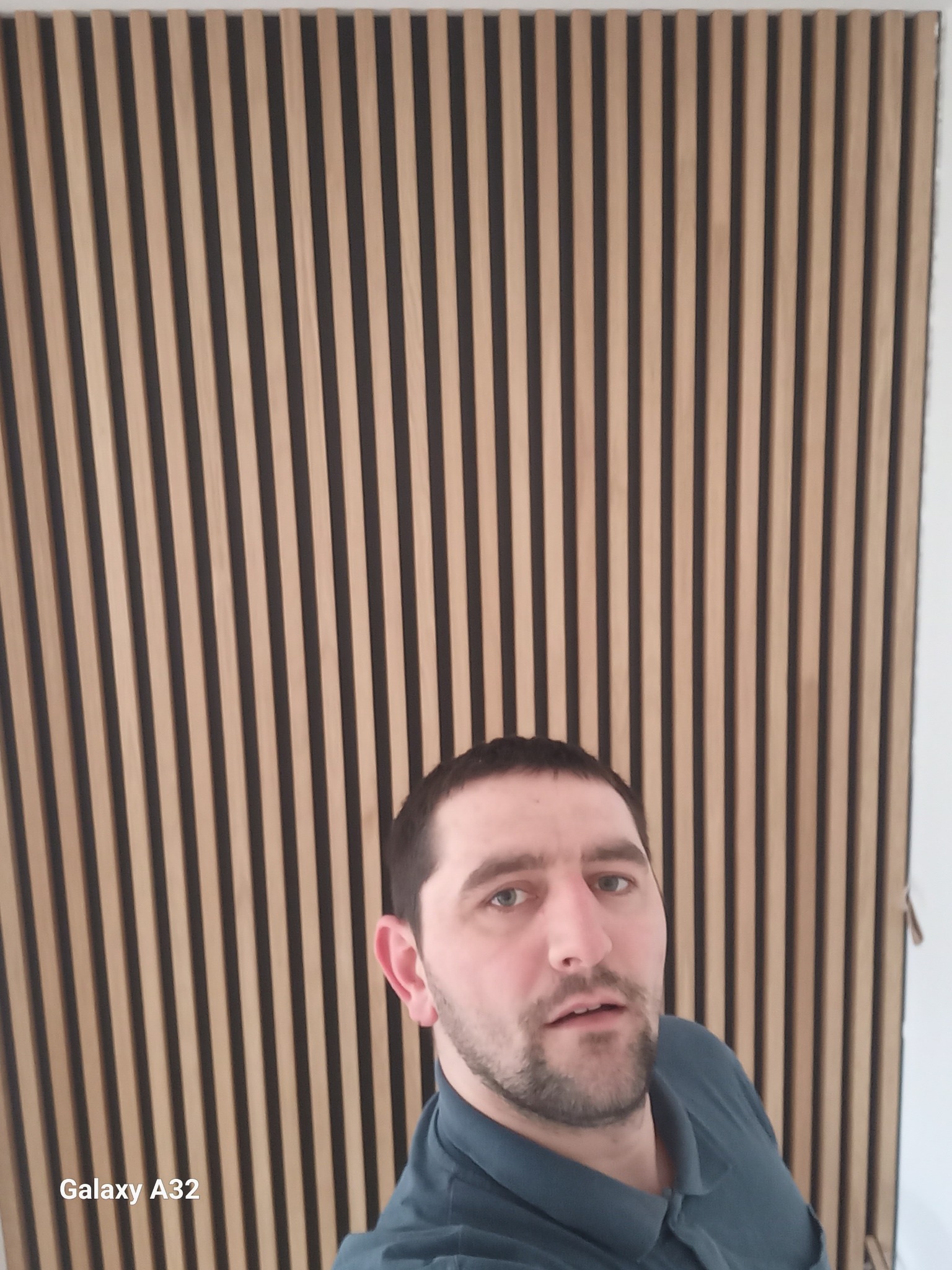 Mężczyzna robi selfie na tle ściany wykończonej panelami dekoracyjnymi z drewna w odcieniach beżu i brązu.