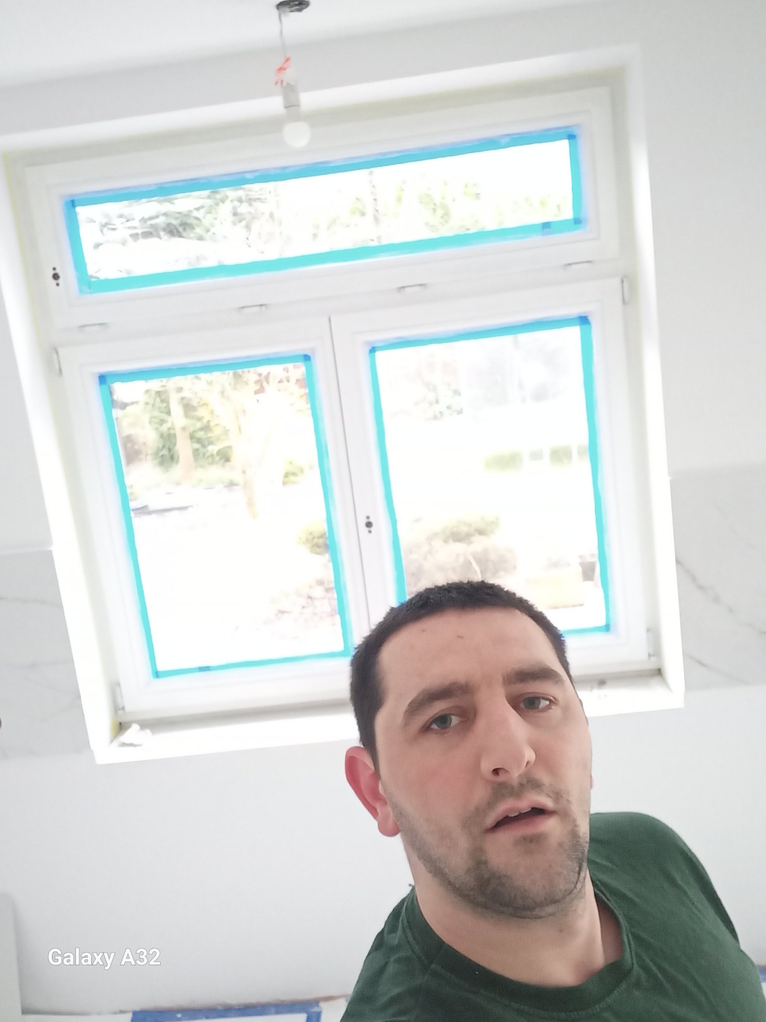 Mężczyzna w zielonej koszulce robi selfie na tle okna dachowego zabezpieczonego taśmą malarską w trakcie prac remontowych w jasnym pomieszczeniu.