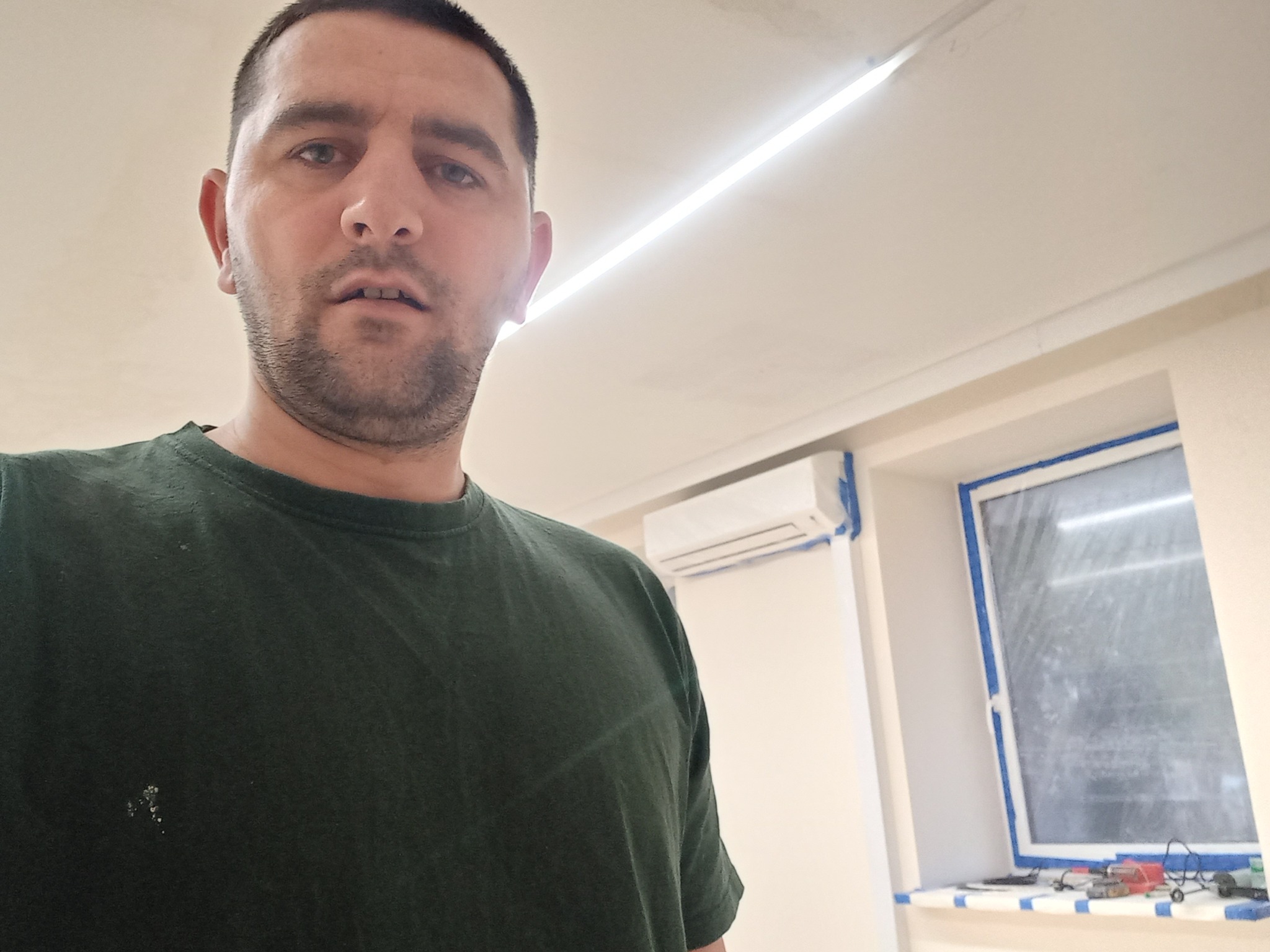 Mężczyzna w zielonej koszulce z plamami farby, robiący selfie w trakcie prac wykończeniowych; widoczne okno zabezpieczone taśmą malarską, klimatyzator i narzędzia na parapecie.