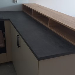 Marek Prządka - Fragment kuchni z ciemnym blatem, jasnymi szafkami i drewnianą półką nad blatem. Na ścianie gniazdko elektryczne i wisząca roślina w metalowej ramie.