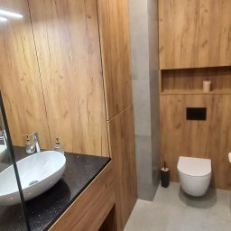 Marek Prządka - Nowoczesna łazienka z drewnianymi szafkami, czarnym blatem pod umywalką nablatową, szarą podłogą i toaletą w zabudowie.