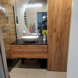 Marek Prządka - Nowoczesna łazienka w Pruszczu Gdańskim z okrągłym lustrem, umywalką nablatową i drewnianą zabudową meblową, widok zza drzwi.