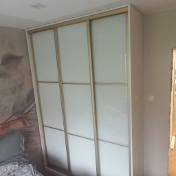 Marek Prządka - Wnętrze sypialni z białą szafą przesuwną z trzema matowymi szklanymi panelami w jasnych drewnianych ramach, widoczna część łóżka z szarą pościelą w kwiaty, na ścianie fototapeta z motywem roślinnym...