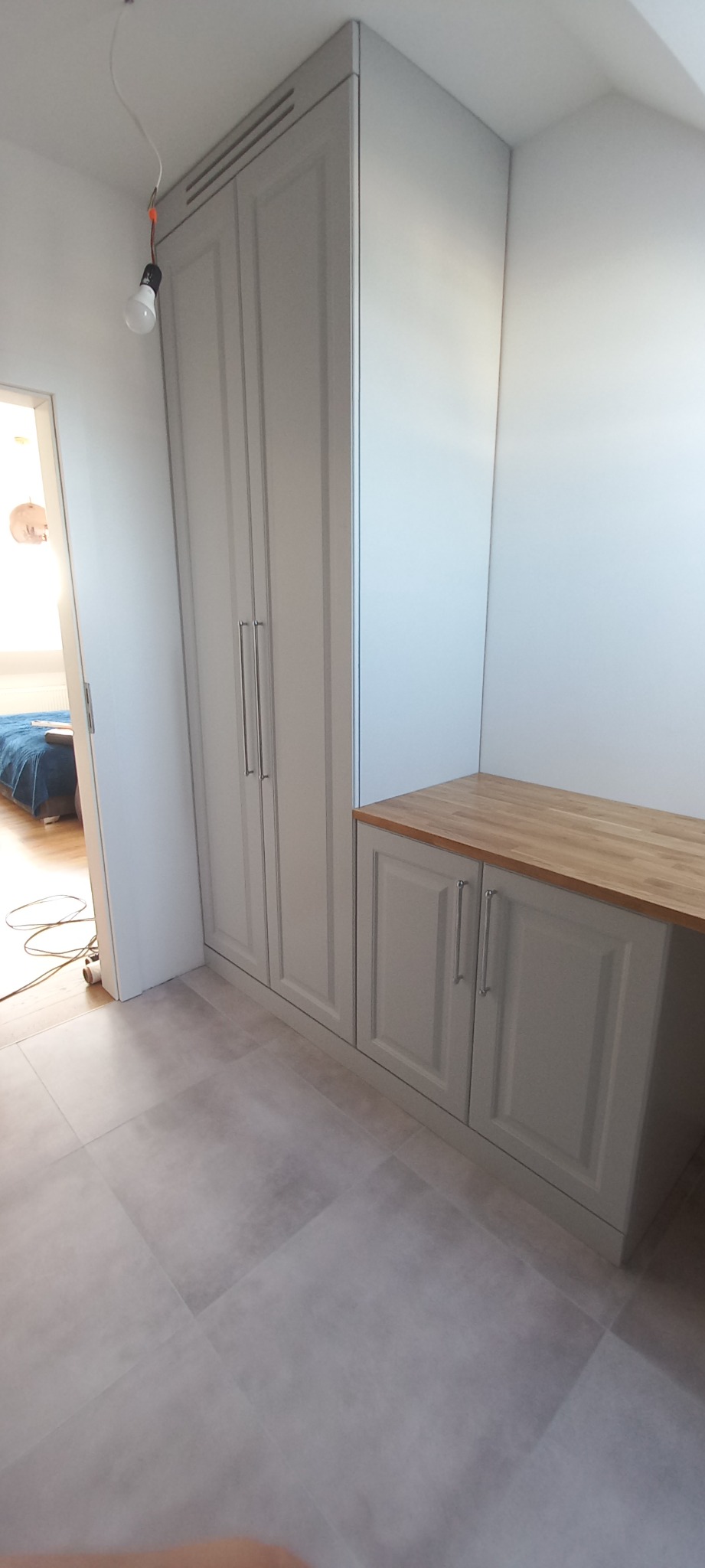 Jasnoszara zabudowa meblowa z szafą dwudrzwiową i szafkami z drewnianym blatem, widok z boku, surowa żarówka zwisająca z sufitu.