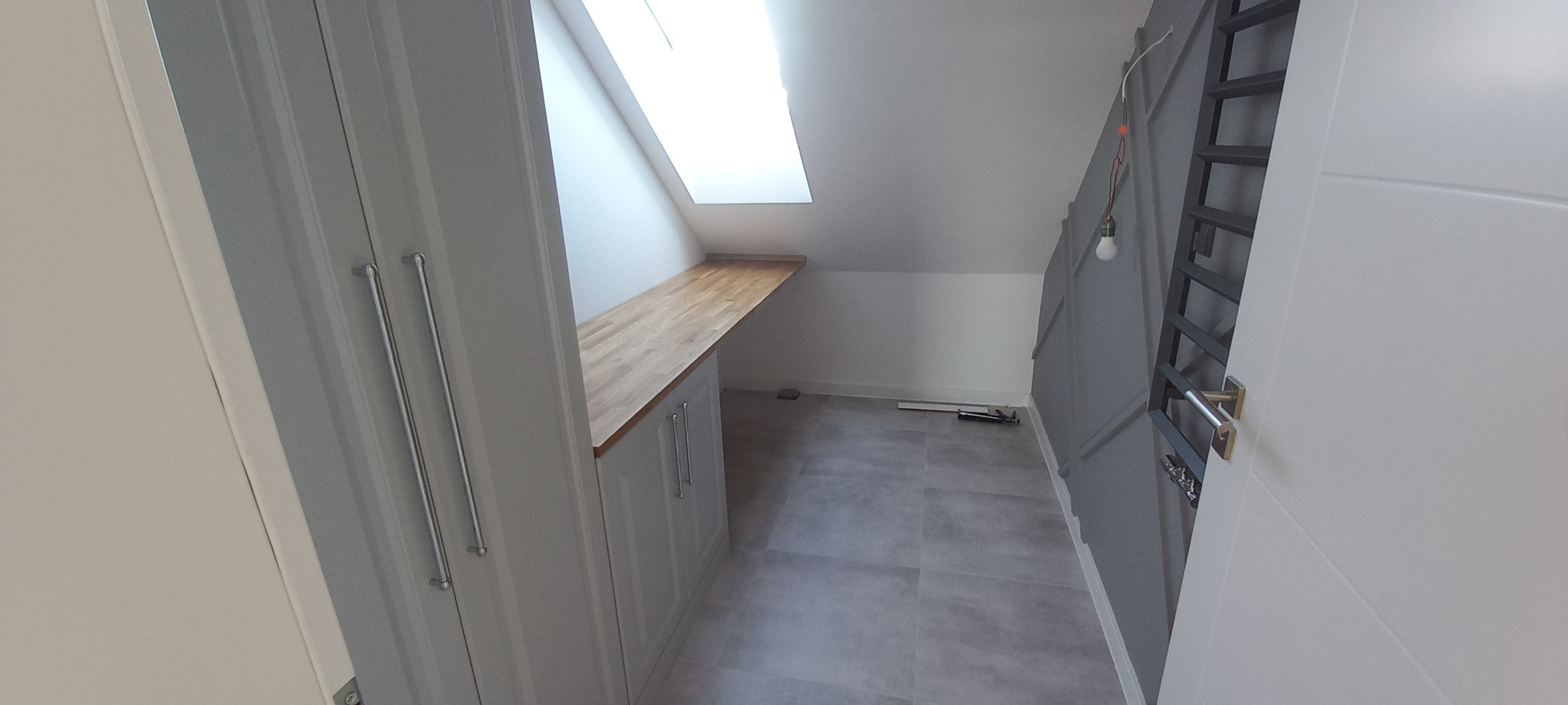 Wnętrze poddasza z zabudową szafy z drewnianym blatem pod oknem dachowym, szara podłoga, grzejnik drabinkowy na ścianie i widoczne narzędzia na podłodze.