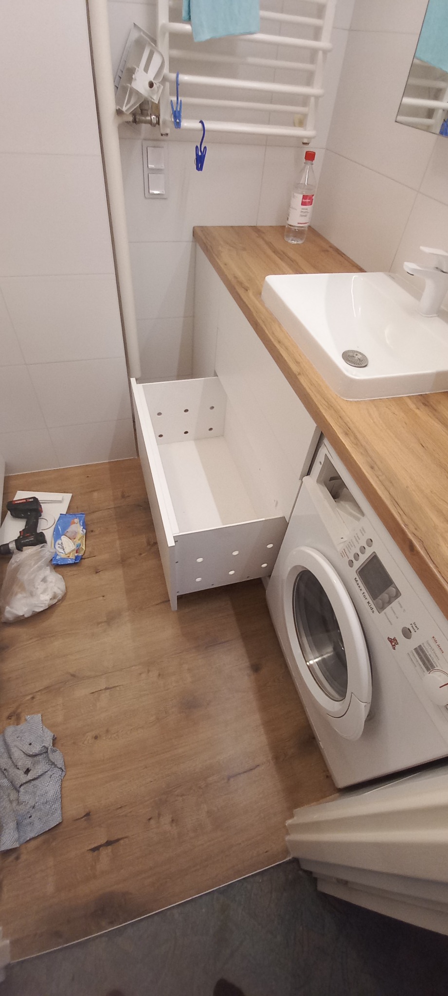 Łazienka w trakcie montażu: wysunięta szuflada pod umywalką z blatem drewnopodobnym, obok pralka, na podłodze narzędzia i resztki opakowań.