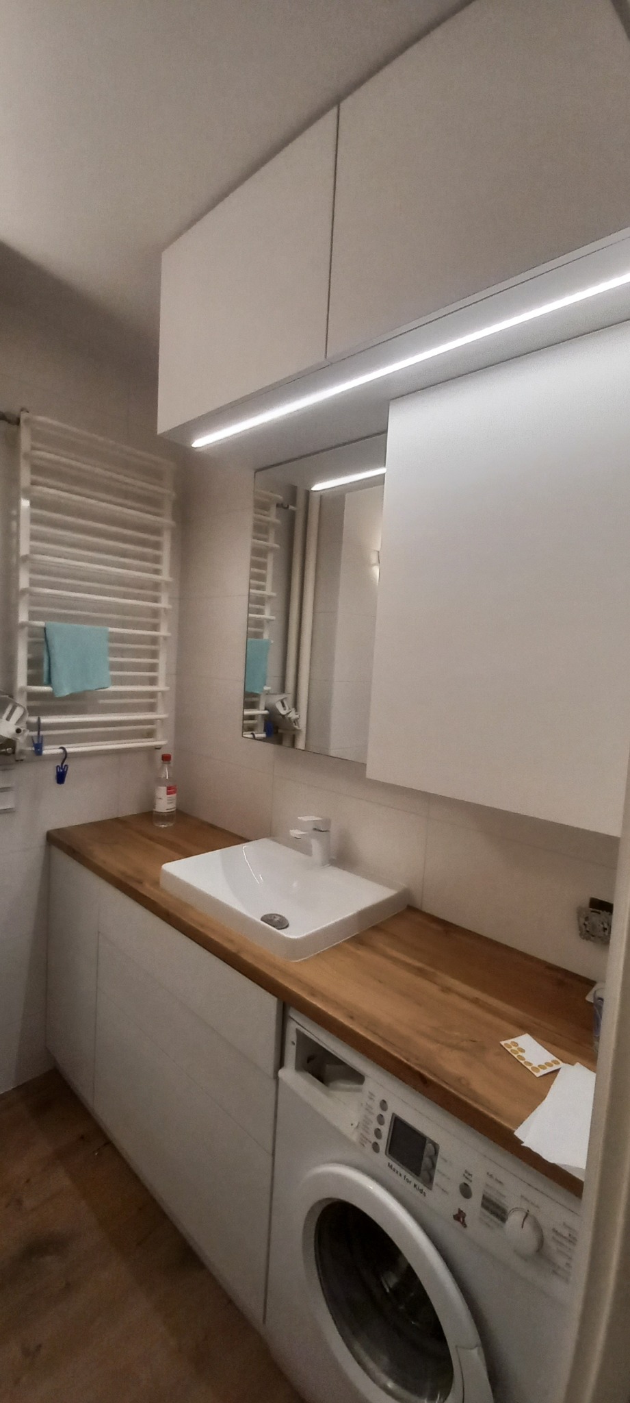 Nowoczesna łazienka z zabudową meblową: białe szafki wiszące, drewniany blat z umywalką nablatową i pralką w zabudowie.