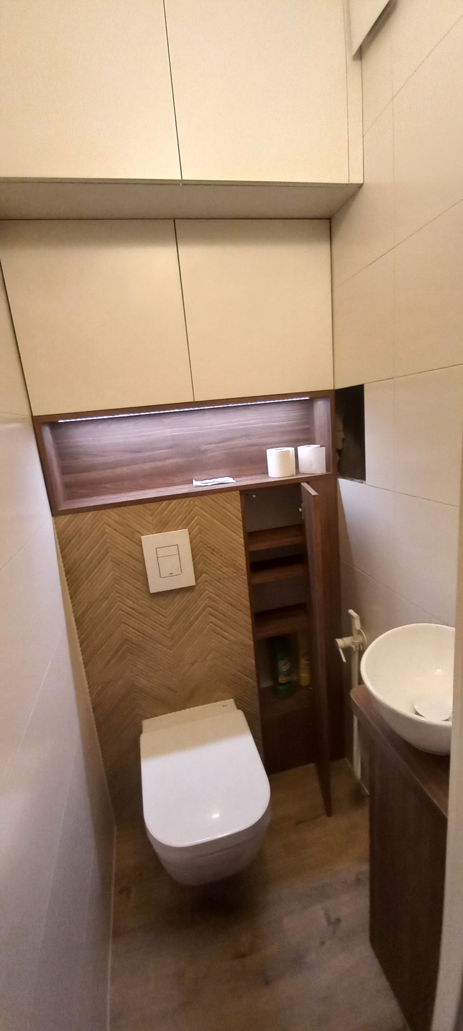 Nowoczesna toaleta z zabudową meblową: szafki wiszące, półki z oświetleniem LED, ściana wykończona drewnopodobną mozaiką w jodełkę, umywalka nablatowa.