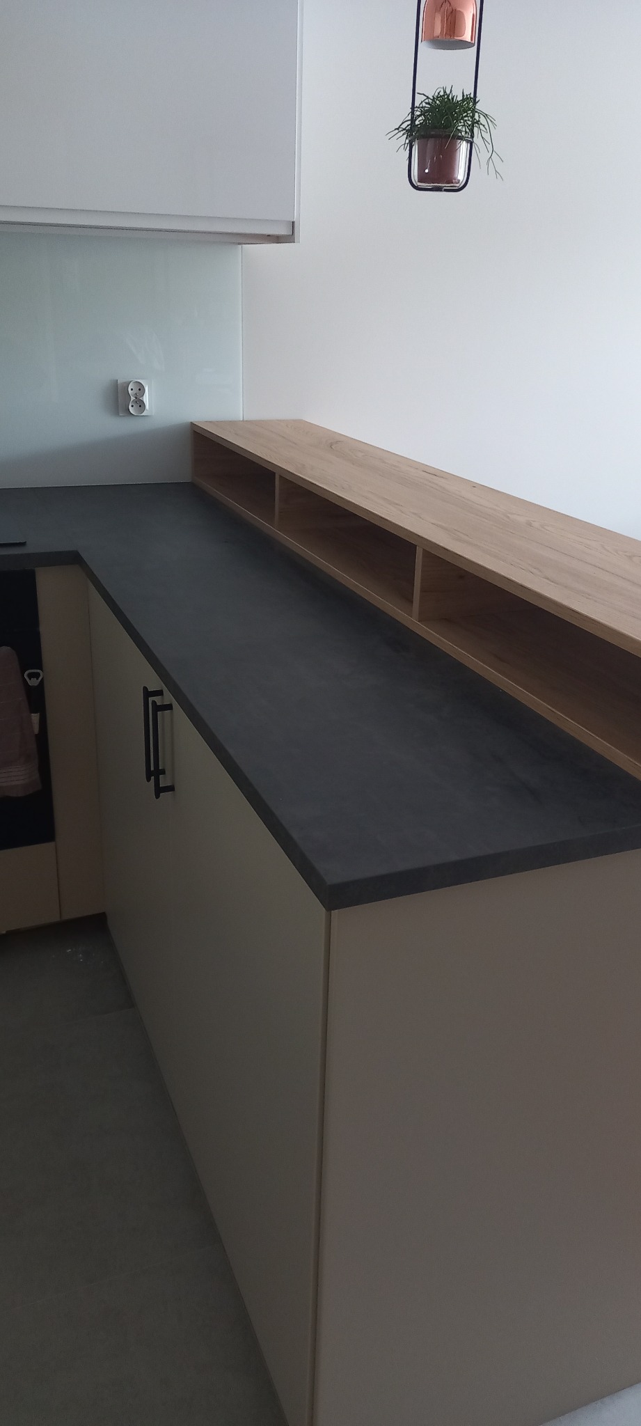Fragment kuchni z ciemnym blatem, jasnymi szafkami i drewnianą półką nad blatem. Na ścianie gniazdko elektryczne i wisząca roślina w metalowej ramie.