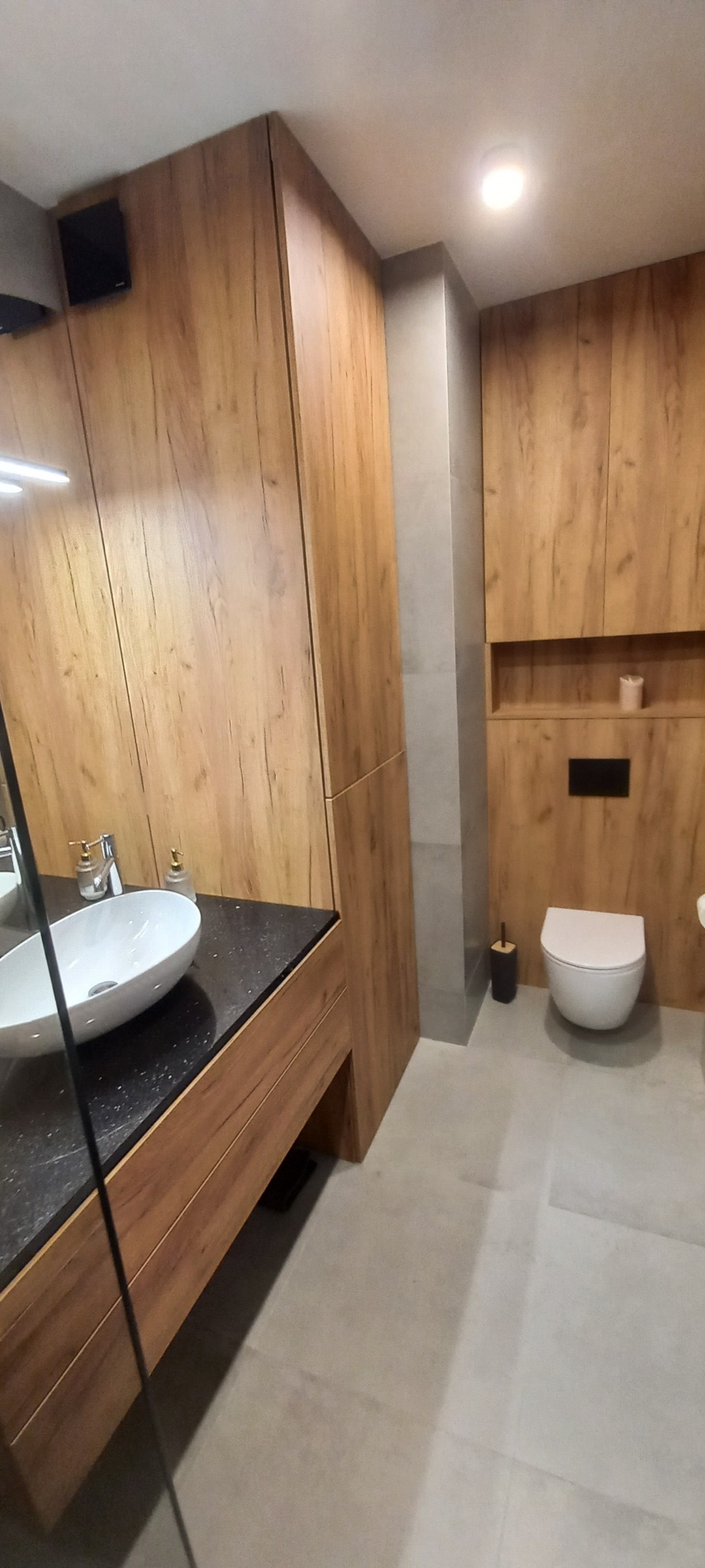 Nowoczesna łazienka z drewnianymi szafkami, czarnym blatem pod umywalką nablatową, szarą podłogą i toaletą w zabudowie.
