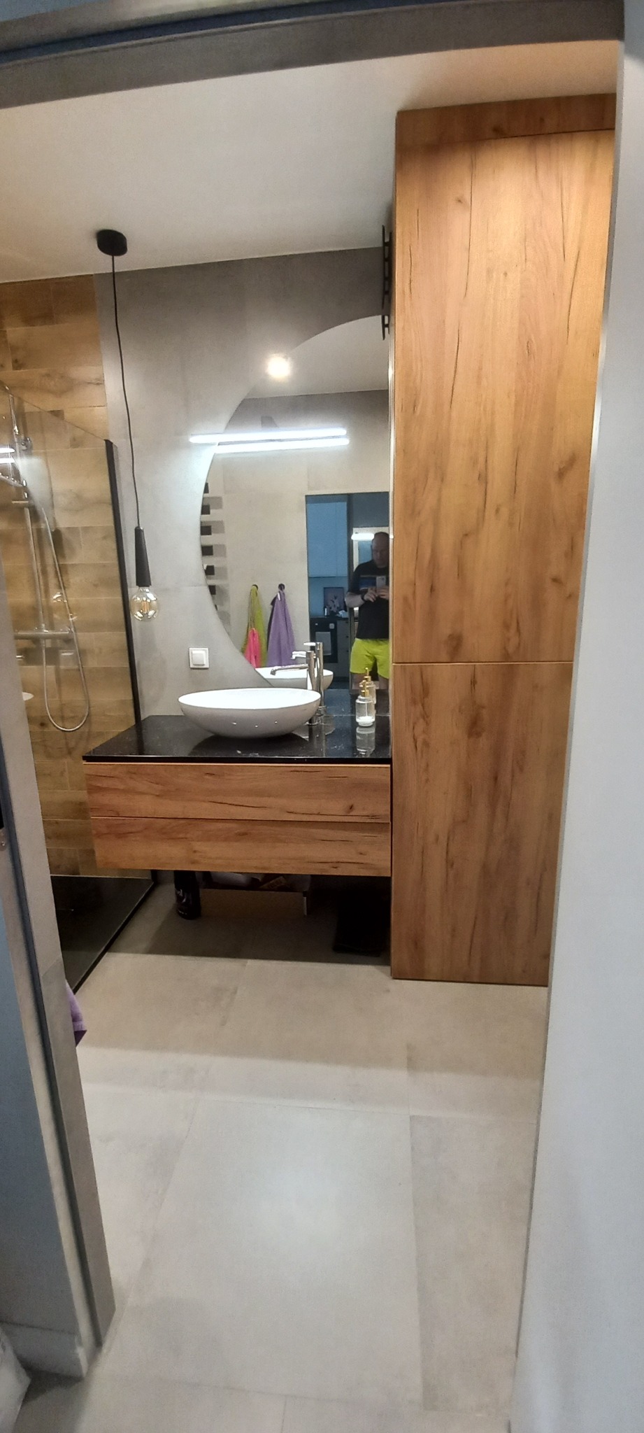 Nowoczesna łazienka w Pruszczu Gdańskim z okrągłym lustrem, umywalką nablatową i drewnianą zabudową meblową, widok zza drzwi.