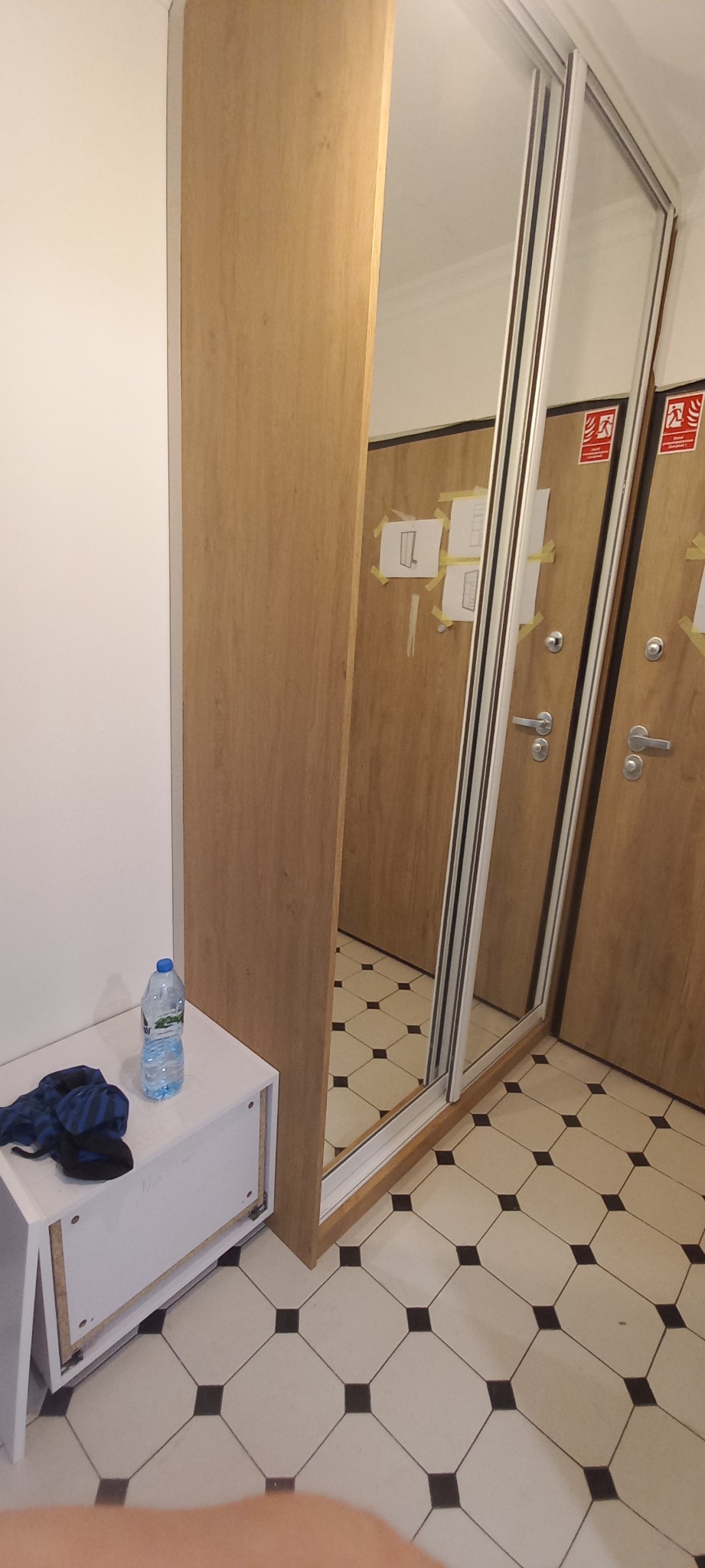 Wnętrze z szafą wnękową z lustrzanymi drzwiami przesuwnymi, obok biała szafka z butelką wody i ubraniem, na podłodze płytki w czarno-białą szachownicę.