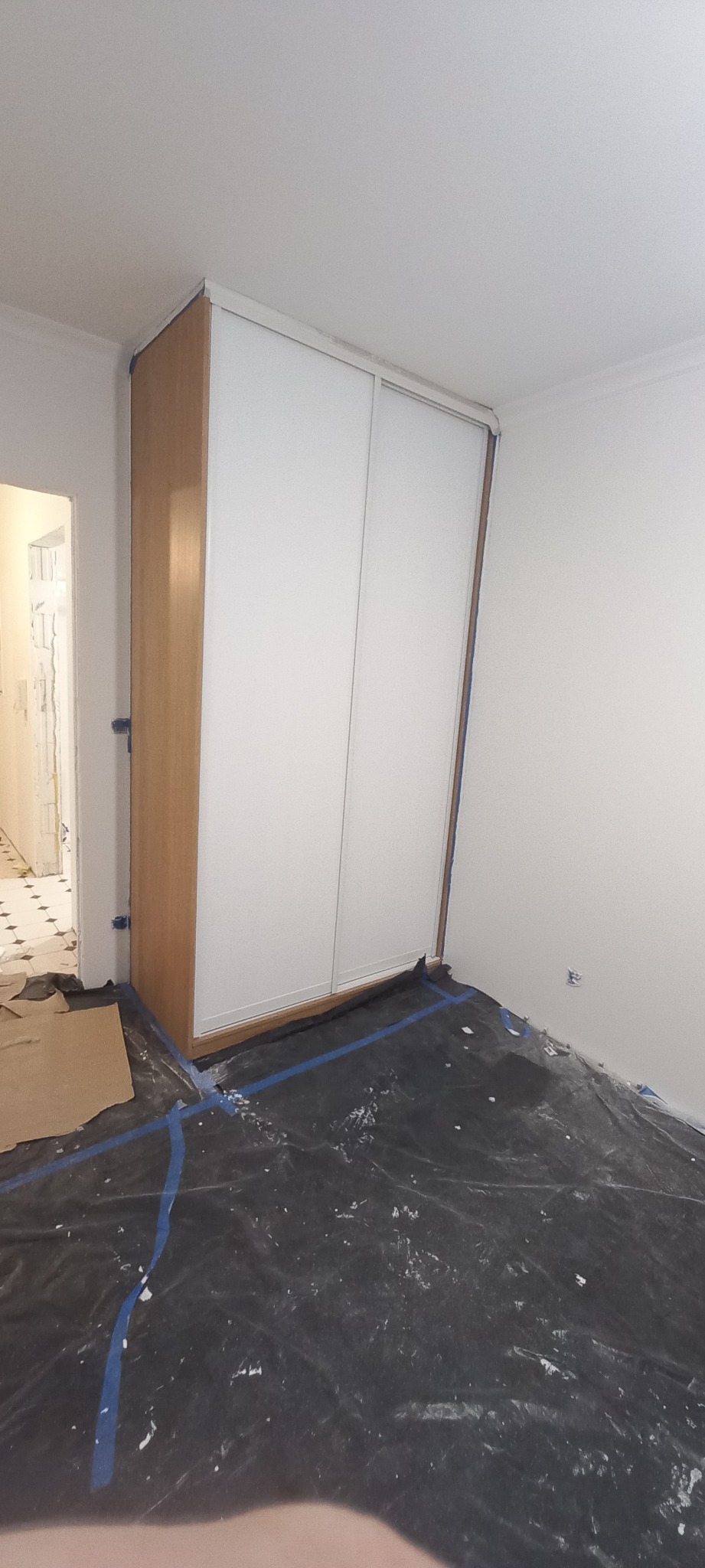 Montaż szafy wnękowej z przesuwnymi, białymi drzwiami; podłoga zabezpieczona folią malarską, widoczne ślady farby na folii i taśma malarska.