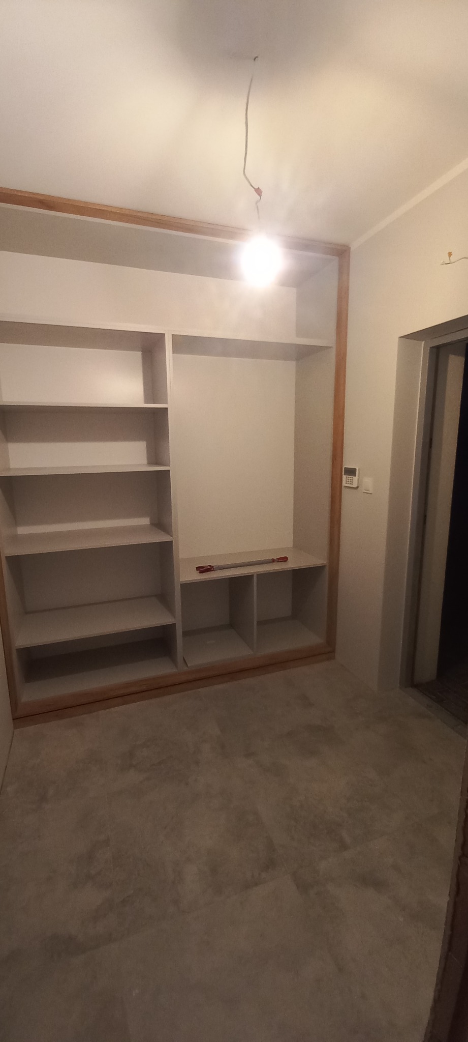 Wnętrze pokoju z wnęką przygotowaną pod zabudowę szafy, widoczne półki i narzędzia na jednej z nich.